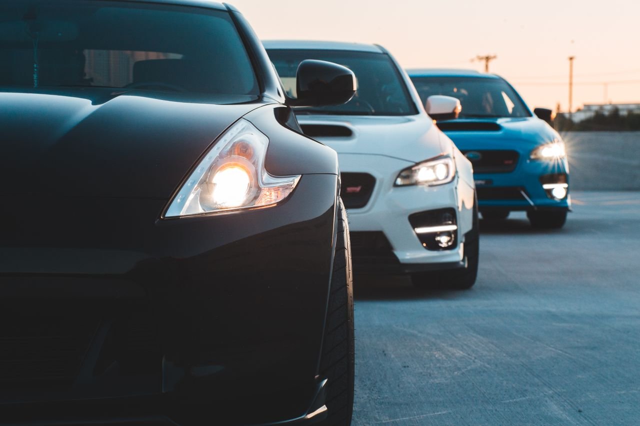 Contemporary sports cars with switched headlights parked in row on asphalt parking in twilight
